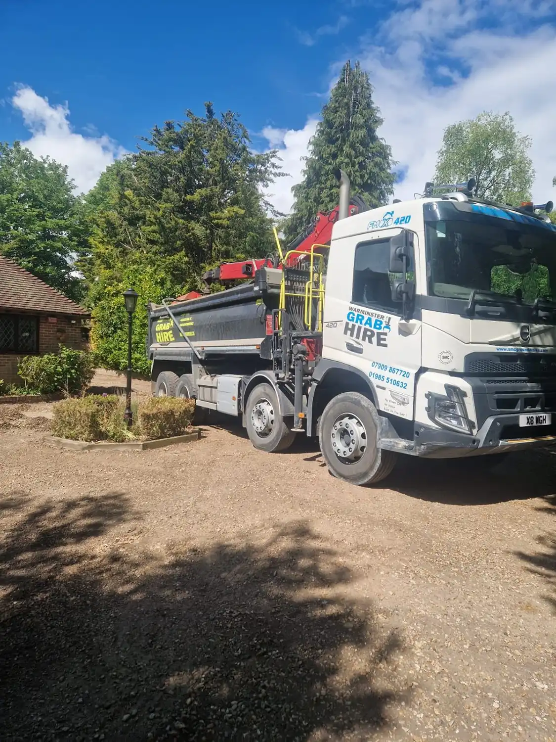 White and black grab hire lorry parked on a gravel driveway near trees and a brick house.