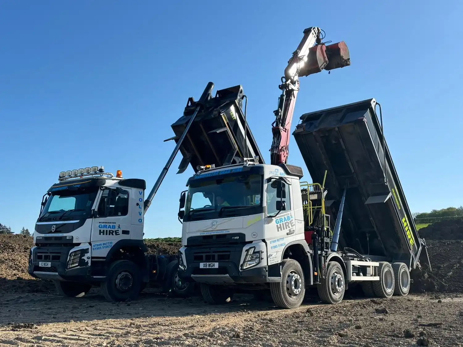 Two white Volvo dump lorries with raised beds at a construction site under blue sky.